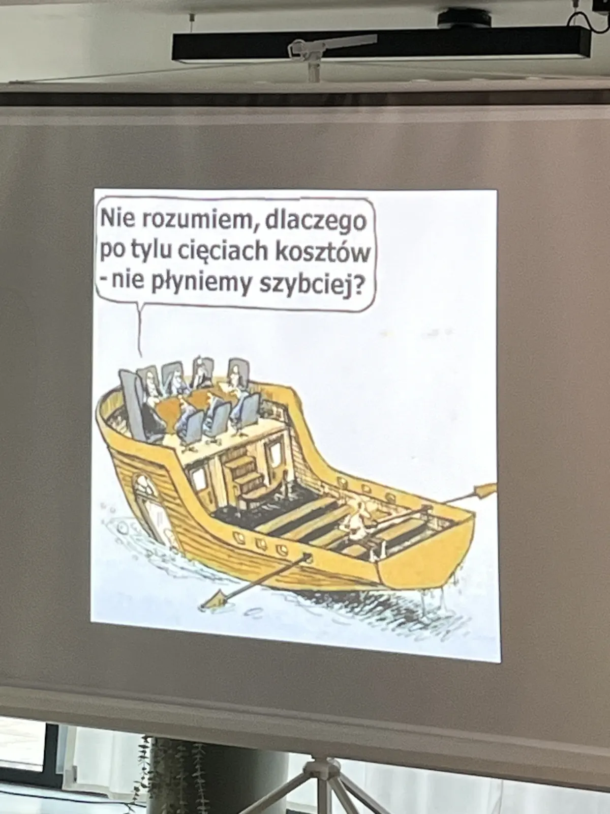 Slajd z WordCamp Wrocław 2026 – rysunek o cięciu kosztów: łódź z jednym wioślarzem i pytaniem, dlaczego nie płyniemy szybciej